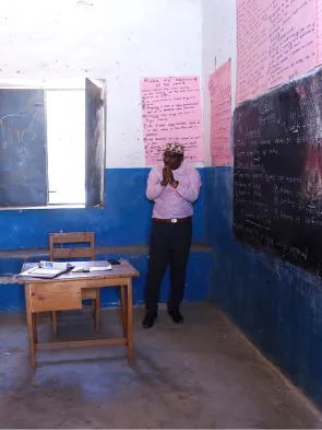 A classroom setting: a teacher in front of a chalkboard talking to a row of girls at desks