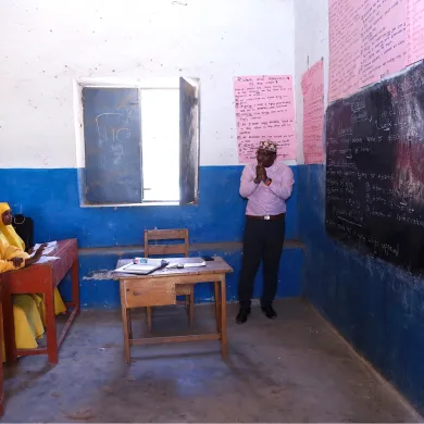 A classroom setting: a teacher in front of a chalkboard talking to a row of girls at desks