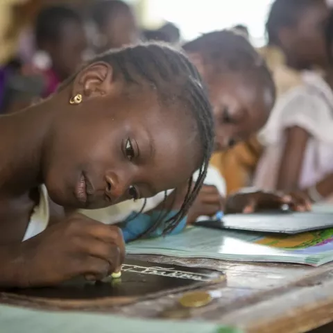 Young girl sitting at a desk and writing on a miniature chalkboard with a piece of chalk