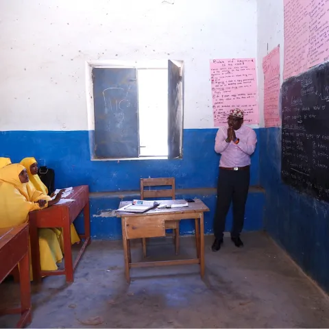 A classroom setting: a teacher in front of a chalkboard talking to a row of girls at desks